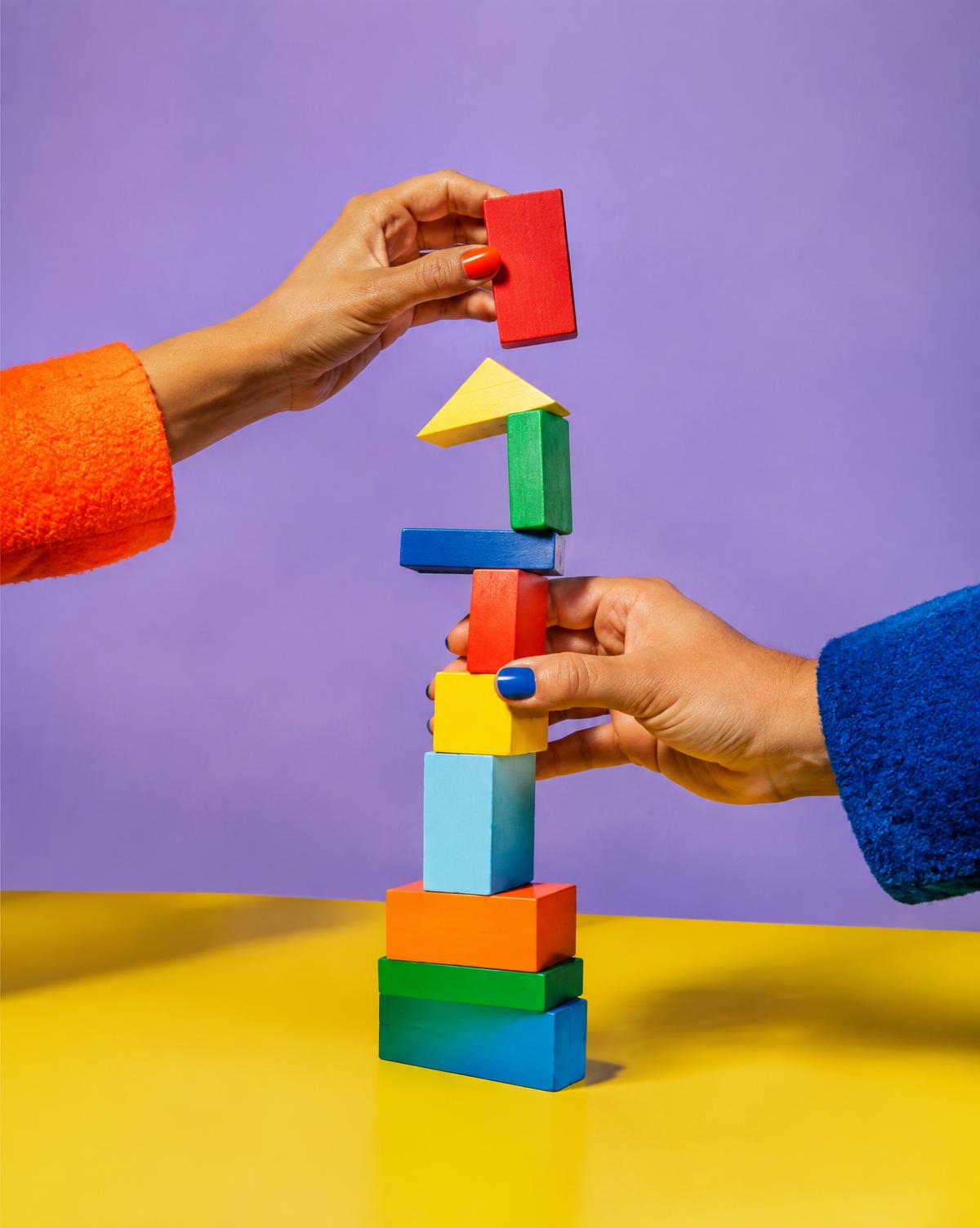 Two hands in orange and cobalt sleeves building a colourful block tower together