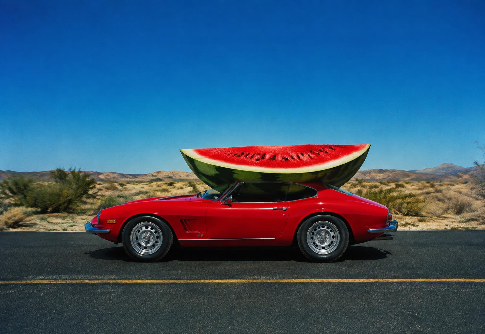 A red sports car on a desert road with a giant slice of watermelon as its roof